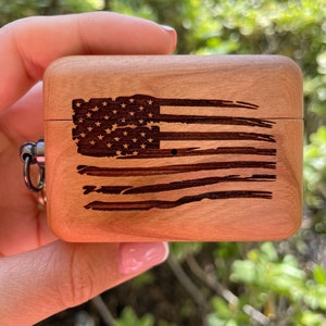 Pode incluir: Uma caixa de madeira clara com um desenho gravado de uma bandeira americana desgastada. A caixa retangular tem bordas arredondadas e um anel de metal prateado. O design da bandeira inclui estrelas e listras.