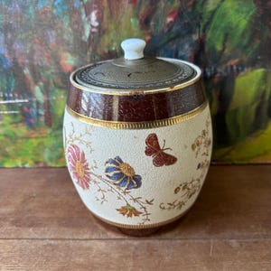 May include: An antique ceramic jar with a lid. The jar is cream-colored with floral and butterfly designs in pink, blue, and brown. It has a dark brown band around the middle and a gold trim. The lid is metal with a white knob.