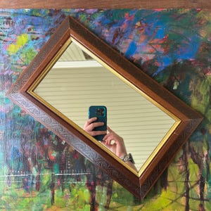 May include: A rectangular mirror with a brown wooden frame and a gold inner border. The frame has a decorative embossed pattern. The mirror reflects a person holding a phone. The background is a colorful abstract painting.