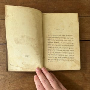 May include: Open antique book with aged, yellowed pages. The text on the right page reads "PREFACE." The book is open, revealing printed text. The book's cover is visible on the left side. The book is on a wooden surface.