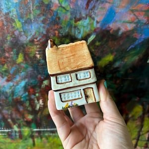 May include: A small ceramic house figurine featuring a tan-colored roof, white walls, and brown trim. The structure includes two upper windows, one lower window, and a single door. A small painted garden with yellow and purple flowers sits at the base.