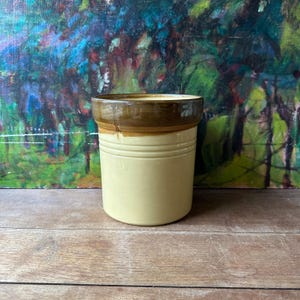 May include: A cylindrical ceramic crock featuring a smooth, light cream-colored body and a contrasting dark brown glazed rim. The vessel has a wide, open top and two horizontal embossed rings encircling the upper portion of the exterior wall.