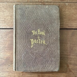 May include: An antique book titled "The Book of Poetry" with gold lettering on the cover. The cover is a muted brown color with a diamond pattern and an oval design around the title. The book appears to be well-worn.