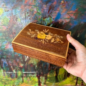 May include: A rectangular wooden box featuring a polished dark brown finish and a light wood inlay border. The lid is decorated with a central floral marquetry design composed of various wood tones, showcasing yellow, tan, and dark brown petals.