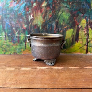 May include: A small, round, metallic pot with a weathered bronze finish, featuring two side handles and three ornate, decorative feet. The vessel has a wide opening and a slightly tapered body, showing signs of patina and surface texture.