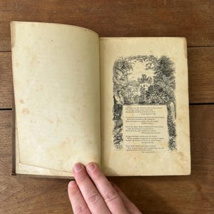 May include: An open antique book with aged, yellowed pages. The right page features an illustrated poem with a detailed border of foliage and figures. The left page is blank. The book is held open by a hand.