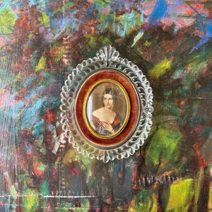 May include: A small, oval portrait of a woman in a decorative, clear plastic frame. The portrait is set against a red velvet background and a gold-colored frame. The woman is wearing a red and blue dress.