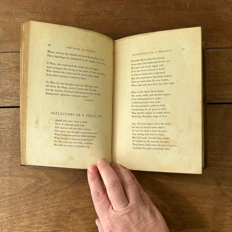 May include: An open antique book with aged, yellowed pages. The left page features the title "REFLECTIONS ON A SKELETON" and poetry. The right page continues the poem. The book is held open by a hand.