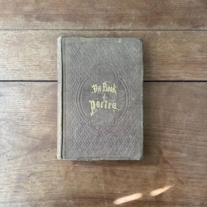 May include: An antique book titled "The Book of Poetry" with gold lettering on a textured, gray cover. The book is closed and resting on a wooden surface. The cover features an ornate, embossed design.