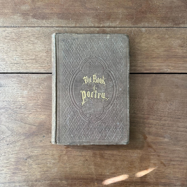 May include: An antique book titled "The Book of Poetry" with gold lettering on a textured, gray cover. The book is closed and resting on a wooden surface. The cover features an ornate, embossed design.
