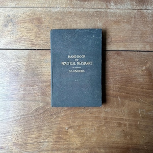 May include: A dark gray hardcover book titled Hand Book of Practical Mechanics by Saunders. The front cover features gold-colored text centered on the surface, displaying the title and author name in a clear, sans-serif font style.