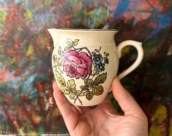 May include: Cream-colored ceramic pitcher with a curved handle and spout. The pitcher is decorated with a large pink rose, blue flowers, and green leaves. The rim is outlined in black.