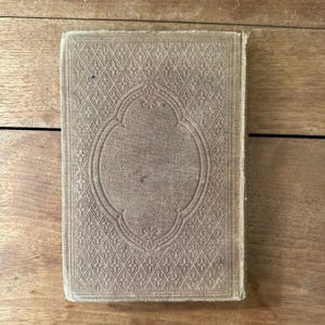 May include: A vintage book with a textured, light brown cover. The cover features an embossed geometric pattern with a central oval design. The book's edges show signs of wear, suggesting its age and use.