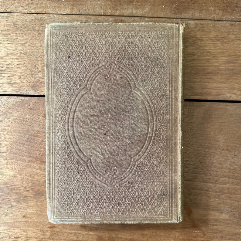 May include: A vintage book with a textured, light brown cover. The cover features an embossed geometric pattern with a central oval design. The book's edges show signs of wear, suggesting its age and use.