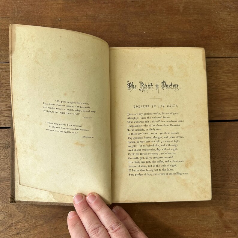 May include: An open antique book with aged, yellowed pages. The right page features the title "The Book of Poetry" and the text "Address to the Deity." The left page has a poem. The book is open on a wooden surface.