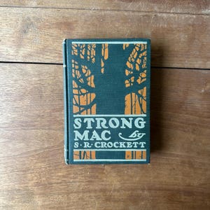 May include: A vintage hardcover book titled "STRONG MAC" by S.R. Crockett. The book has a dark green cover with an orange tree design and white lettering. The book is on a wooden surface.