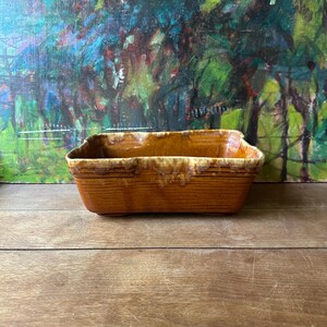 May include: A rectangular ceramic baking dish with a glossy amber-brown glaze. The exterior features horizontal ribbed detailing along the upper rim, while the top edge displays a lighter, cream-colored drip glaze effect over the darker base.