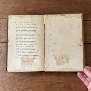 May include: An open antique book with aged, stained pages. The text, titled "THE BOOK OF POETRY," is visible on the left page. The right page shows water stains. The book's cover is a dark brown color. A hand is visible on the right side of the image.