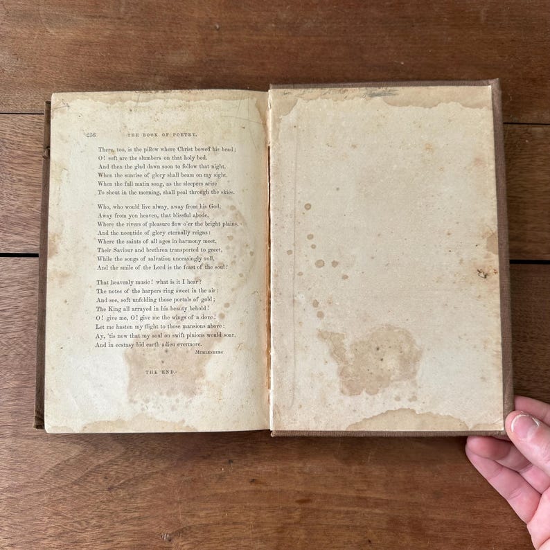 May include: An open antique book with aged, stained pages. The text, titled "THE BOOK OF POETRY," is visible on the left page. The right page shows water stains. The book's cover is a dark brown color. A hand is visible on the right side of the image.