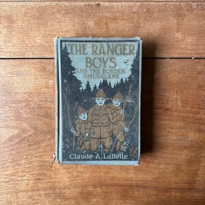 May include: Vintage book titled "The Ranger Boys and the Border Smugglers" by Claude A. LaBelle. The cover features an illustration of three figures in ranger attire, set against a forest backdrop. The book has a distressed, aged appearance.
