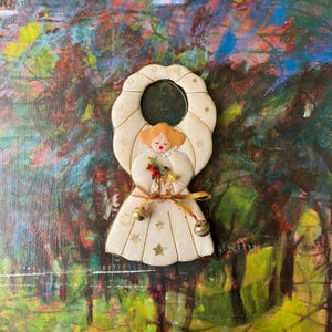 May include: A cream-colored angel Christmas ornament with gold accents. The angel has a halo, wings, and a dress adorned with gold stars. It holds a golden trumpet and a sprig of red berries. Two small bells hang from the bottom.
