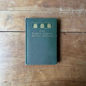 May include: A vintage, dark green book titled "Fifty English Classics Briefly Outlined." The book's cover features gold floral designs and text. The book is rectangular and appears to be in good condition, resting on a wooden surface.
