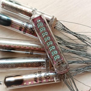 Może przedstawiać: Zbliżenie na grupę zabytkowych lamp Nixie, z jedną lampą wyświetlającą cyfry 00:00:00. Lampy są wykonane z przezroczystego szkła z czerwonym i czarnym wnętrzem. Lampy mają dużą liczbę cienkich przewodów wystających od dołu.