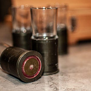 May include: Four clear glass shot glasses with black metal bases. The glasses are arranged in a row on a wooden surface. The glasses are empty.