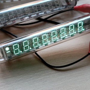 Może przedstawiać: Dwa przezroczyste szklane tubusy Nixie z zielonymi świecącymi cyframi. Tubusy są połączone przewodami i znajdują się na jasnym drewnianym podłożu.
