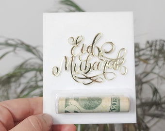 May include: A white card with gold script that reads "Eid Mubarak" and a rolled-up bill. The card is held in front of a plant. The gold script is in a decorative font.