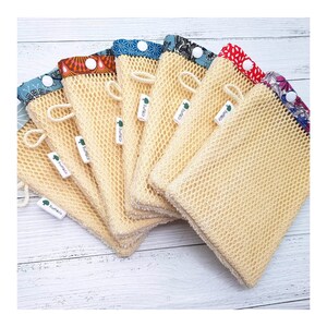 Può includere: Una pila di sei sacchetti per la biancheria in rete beige con bordi in tessuto colorato e una chiusura a bottone bianco. Ogni sacchetto ha un piccolo cartellino verde con la scritta "Turf & Co" stampata su di esso.