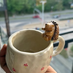 May include: A handmade ceramic mug with a glossy off-white finish and a textured, uneven surface. The mug features small red and white mushroom illustrations and a three-dimensional brown hedgehog figurine attached to the top of the handle.