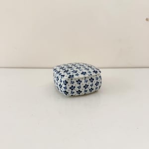 May include: A small, square trinket box with a lid. The box and lid are white with a repeating pattern of small, blue floral designs. The box is on a white surface.