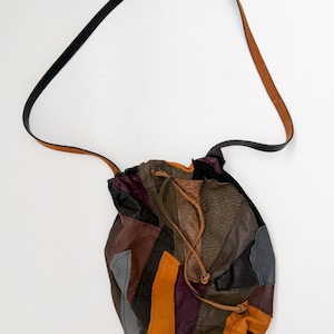 May include: A patchwork leather bucket bag with a long adjustable strap. The bag is made of different coloured leather pieces, including brown, grey, purple, and orange.