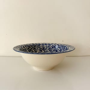 May include: A ceramic bowl with a cream-coloured exterior and a wide, flared rim featuring a dense, intricate blue floral pattern. The interior of the bowl is decorated with the same blue floral design, creating a contrast against the solid cream base.