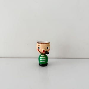 May include: A vintage wooden egg cup in the shape of a man with a painted face, a green body, and a black pipe. The egg cup has a beige head with a red nose and black eyes. The base is black.