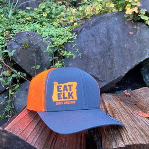 May include: A gray and orange trucker hat with the text "EAT ELK" in orange, along with the outline of the state of Oregon. The hat also includes the text "100% ORGANIC". The hat is sitting on a wooden surface.