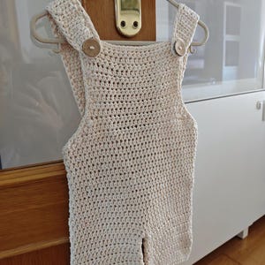 Puede incluir: Un pelele de ganchillo color crema con un patrón de puntos sencillo, dos tirantes y dos botones redondos de madera en la parte delantera. La prenda se muestra en una percha de plástico clara, destacando su textura y su confección.