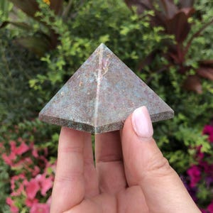 May include: A small, pyramid-shaped stone sculpture with a mottled teal and brown surface. The pyramid is held in a hand against a backdrop of green and pink foliage. The stone has a rough, textured appearance.