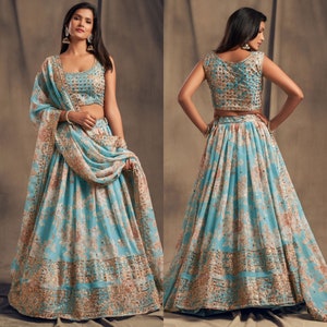 May include: A two-piece turquoise lehenga set with a floral pattern in peach and gold. The sleeveless top and long skirt are adorned with intricate gold embellishments. A matching dupatta drapes over the shoulder, completing the traditional Indian ensemble.