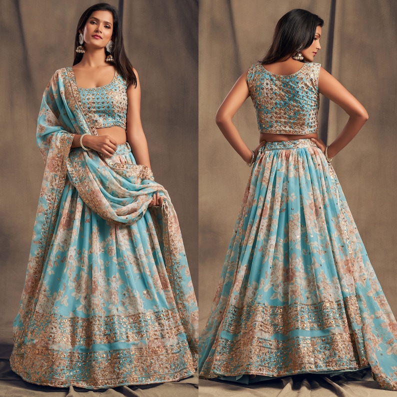 May include: A two-piece turquoise lehenga set with a floral pattern in peach and gold. The sleeveless top and long skirt are adorned with intricate gold embellishments. A matching dupatta drapes over the shoulder, completing the traditional Indian ensemble.