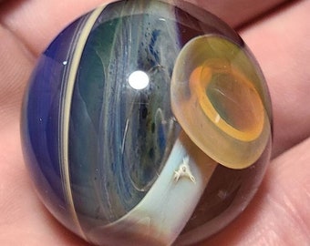 May include: A glass marble with a swirling pattern of blue, white, and yellow. The marble has a clear centre and a ring of orange around the centre.