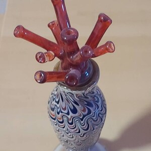 May include: A small, hand-blown glass vase with a swirling black, white, and orange pattern. The vase has a wide, round base and a narrow neck. A cluster of red glass tubes with rounded ends are arranged in the vase, resembling a flower.