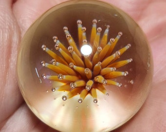 May include: A clear glass marble featuring a central design of yellow and orange spikes, reminiscent of a burst or flower. Tiny air bubbles are visible within the glass, enhancing its unique aesthetic. The marble is a decorative item.