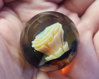 May include: A glass marble with a yellow flower encased inside. The marble has a dark brown, black, and orange swirl pattern.