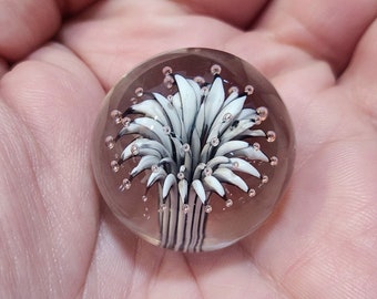 May include: A clear glass paperweight with a white and black flower design inside. The flower has a stem and multiple petals. There are small air bubbles trapped inside the glass.