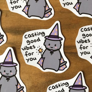 May include: A set of eight stickers featuring a cartoon grey cat wearing a purple and yellow wizard hat. The cat is casting good vibes for you. The stickers are on a wooden surface.