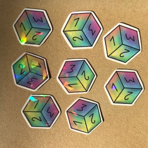 May include: Nine iridescent holographic stickers of a six-sided die with numbers 1 through 6. The stickers are arranged in a circle on a brown surface.