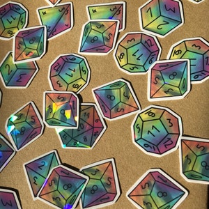 Puede incluir: Una colección de pegatinas holográficas de arcoíris iridiscentes con diferentes números en una superficie marrón. Las pegatinas tienen forma de dados y tienen contornos negros.