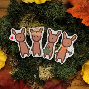 May include: Four brown cartoon animal stickers with different accessories. The stickers are on a green moss background with fall leaves.